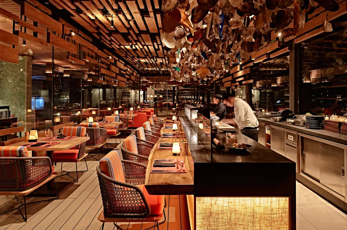 Finolhu Baa Atoll, dining, Baa Atoll, Maldives, Kanusan Japanese teppanyaki restaurant interior, the long counter with the chef working at the live-cooking station, terracotta-and-coral cushioned woven chairs along the wood bench, suspended ceiling installation of carved bowls and shells inspired by the Maldivian sea.