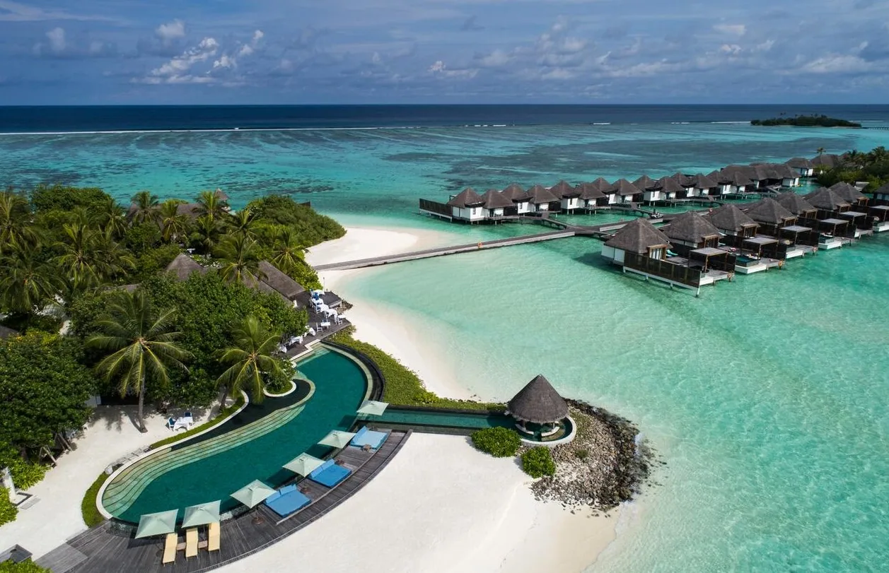 Four Seasons Resort Maldives at Kuda Huraa, aerial, North Malé Atoll, Maldives, Lateral aerial of the resort, the kidney-shaped main pool curving toward the white-sand crescent and the overwater villa row extending east across the reef.