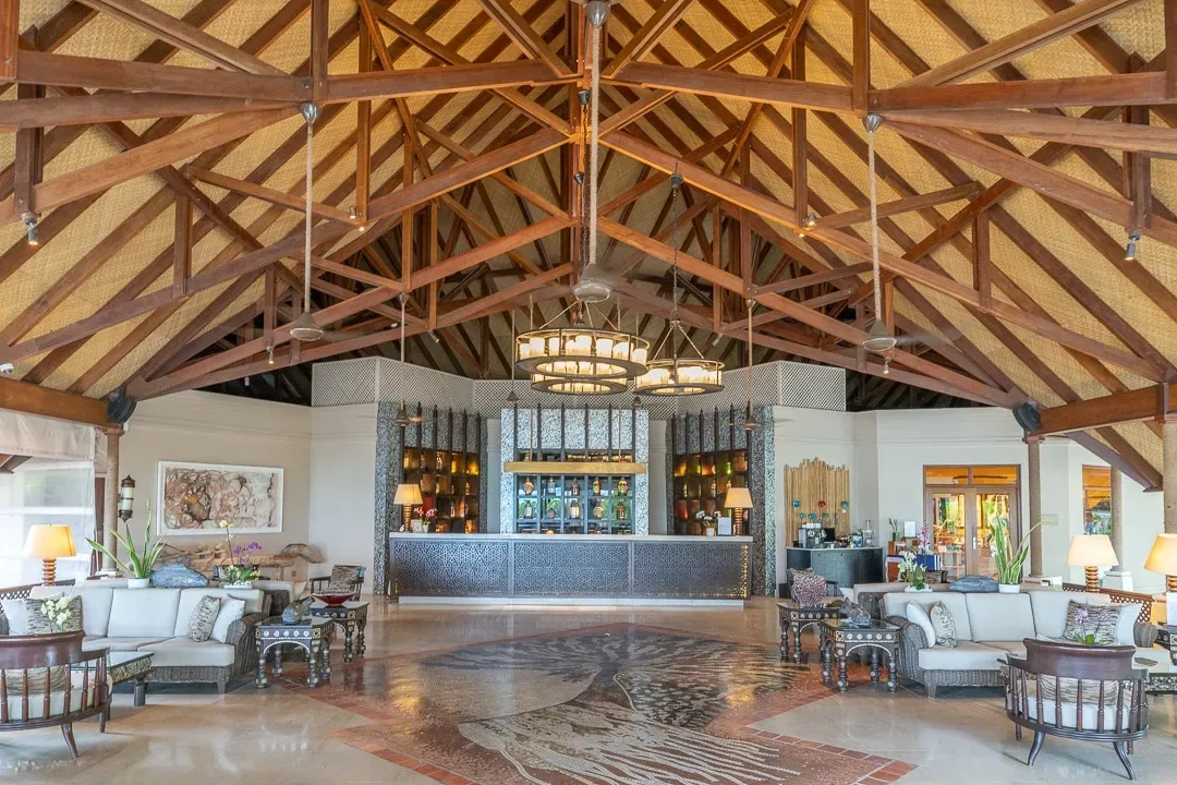 Four Seasons Resort Maldives at Kuda Huraa, arrival, North Malé Atoll, Maldives, Main lobby interior, exposed timber-truss ceiling under a vaulted thatched roof, central reception desk in dark timber with a chandelier above, woven-stone floor pattern and mid-tone rattan armchairs flanking the entry.