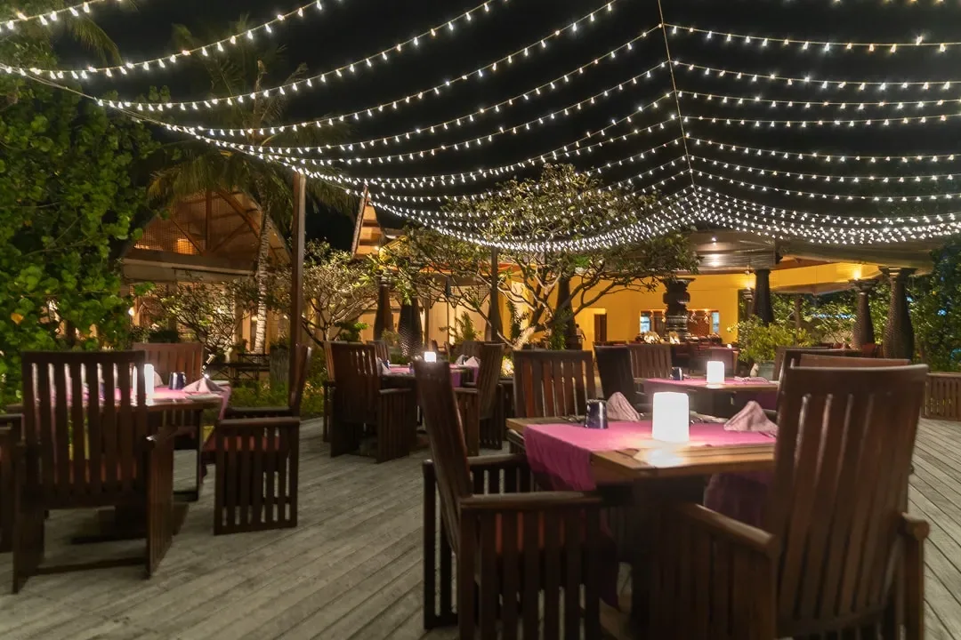 Four Seasons Resort Maldives at Kuda Huraa, dining, North Malé Atoll, Maldives, Baraabaru lagoon-top Indian venue, fairy-light canopy strung between palms over dark-timber dining chairs, magenta linen runners and candle-lamp centrepieces with the lit kitchen pass glowing in the background.