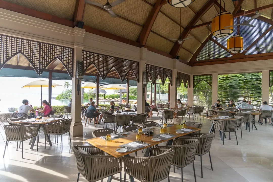 Four Seasons Resort Maldives at Kuda Huraa, dining, North Malé Atoll, Maldives, Café Huraa open-pavilion breakfast service, vaulted ceiling with woven palm panels and dark timber beams, blonde-rattan dining chairs around timber tables, the lagoon visible past the eaves.