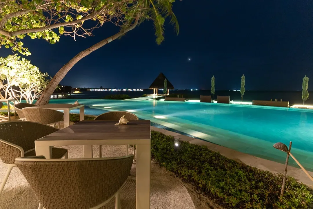 Four Seasons Resort Maldives at Kuda Huraa, dining, North Malé Atoll, Maldives, Reef Club at night, woven dining chairs and timber tables on the sand, the floodlit infinity pool stretching to the dark horizon and the thatched pavilion silhouetted in the distance.