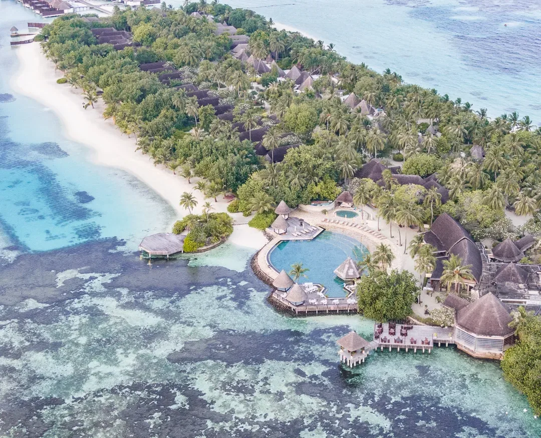 Four Seasons Resort Maldives at Kuda Huraa, setting, North Malé Atoll, Maldives, Top-down aerial of the central island, the main pool ring and thatched dining pavilions clustered above the reef edge, traditional dhonis moored along the inside lagoon.