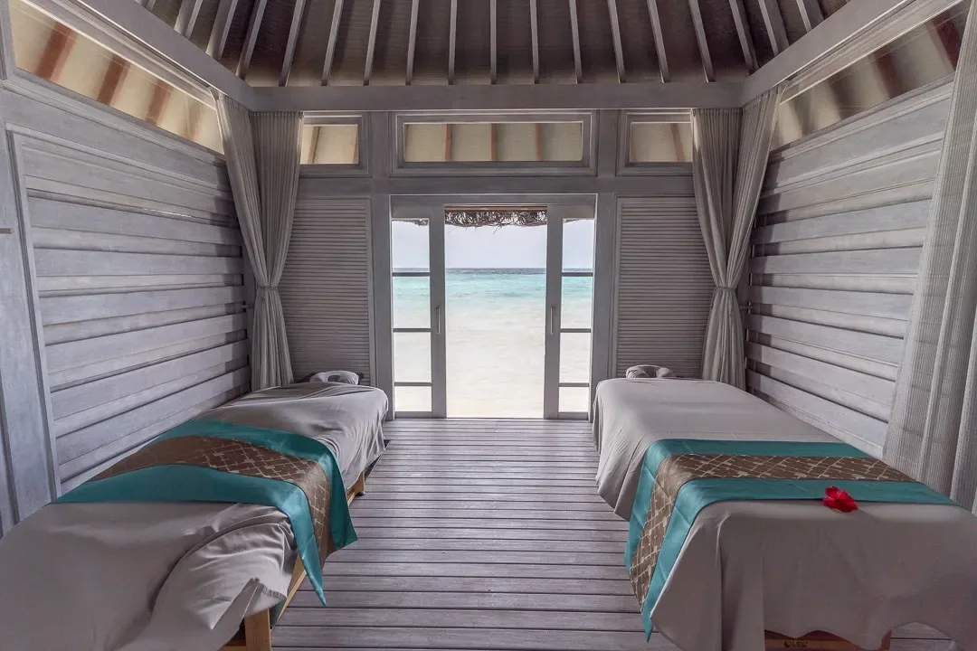 Four Seasons Resort Maldives at Kuda Huraa, spa, North Malé Atoll, Maldives, Island Spa twin treatment room, painted-grey louvred walls and pitched timber ceiling, two massage beds dressed in white linen with teal-and-gold sashes, full-width doors opening directly onto the white-sand beach and the lagoon beyond.