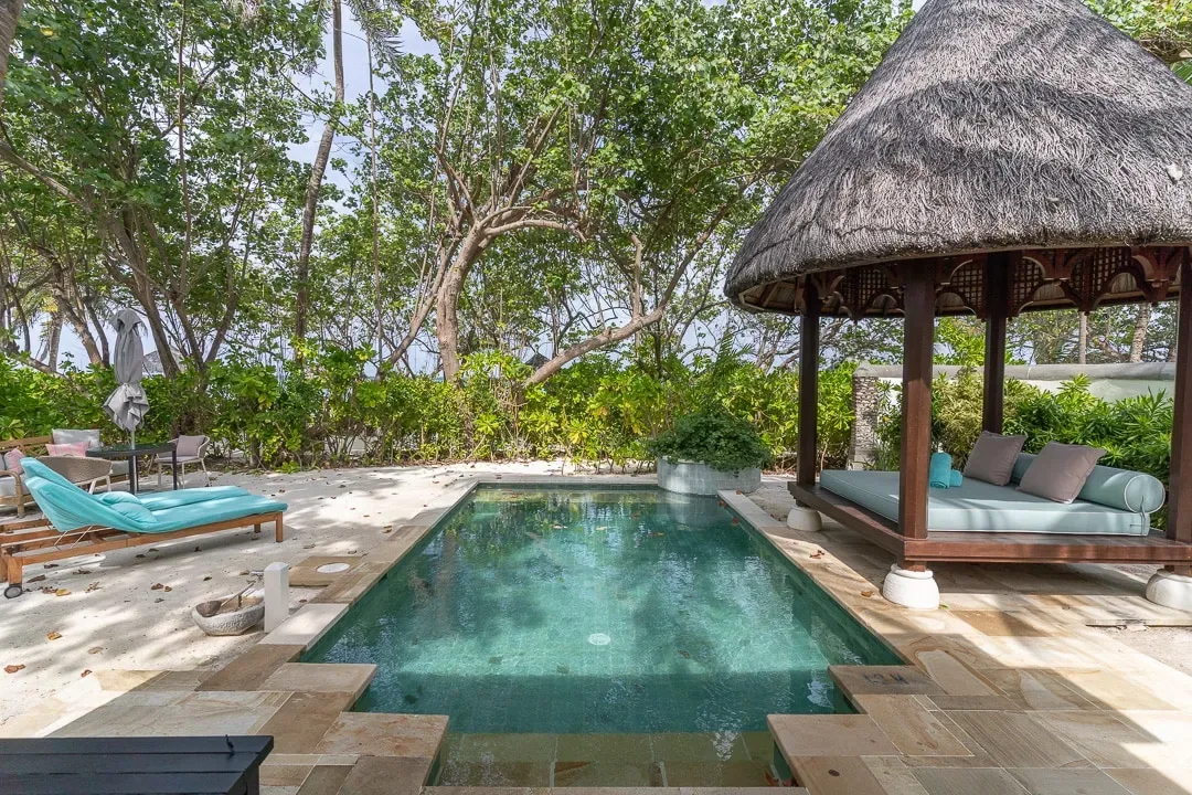 Four Seasons Resort Maldives at Kuda Huraa, villa beach, North Malé Atoll, Maldives, Deluxe Beach Pavilion plunge pool, sandstone deck under tree canopy, thatched daybed gazebo on the right, single sun lounger on the left with the bungalow's jungle backdrop.