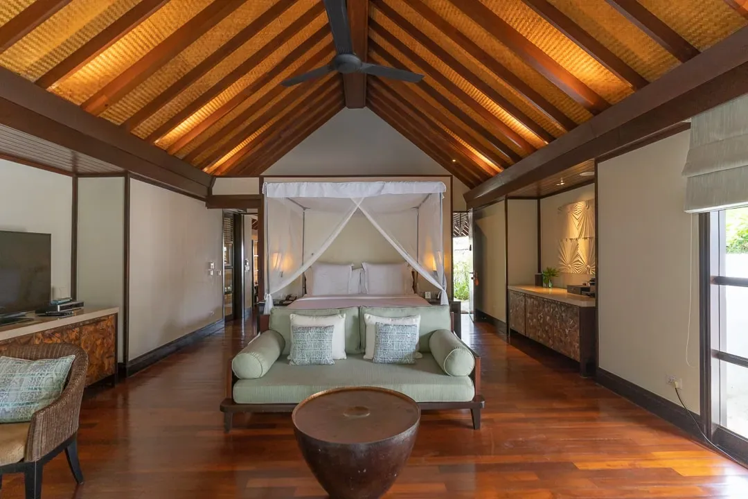 Four Seasons Resort Maldives at Kuda Huraa, villa beach, North Malé Atoll, Maldives, Sunrise Beach Bungalow interior, vaulted thatched ceiling with woven palm panels, four-poster bed under a fine-mesh canopy, dark-hardwood floor, mint-green daybed at the foot, drum coffee table in carved local timber.