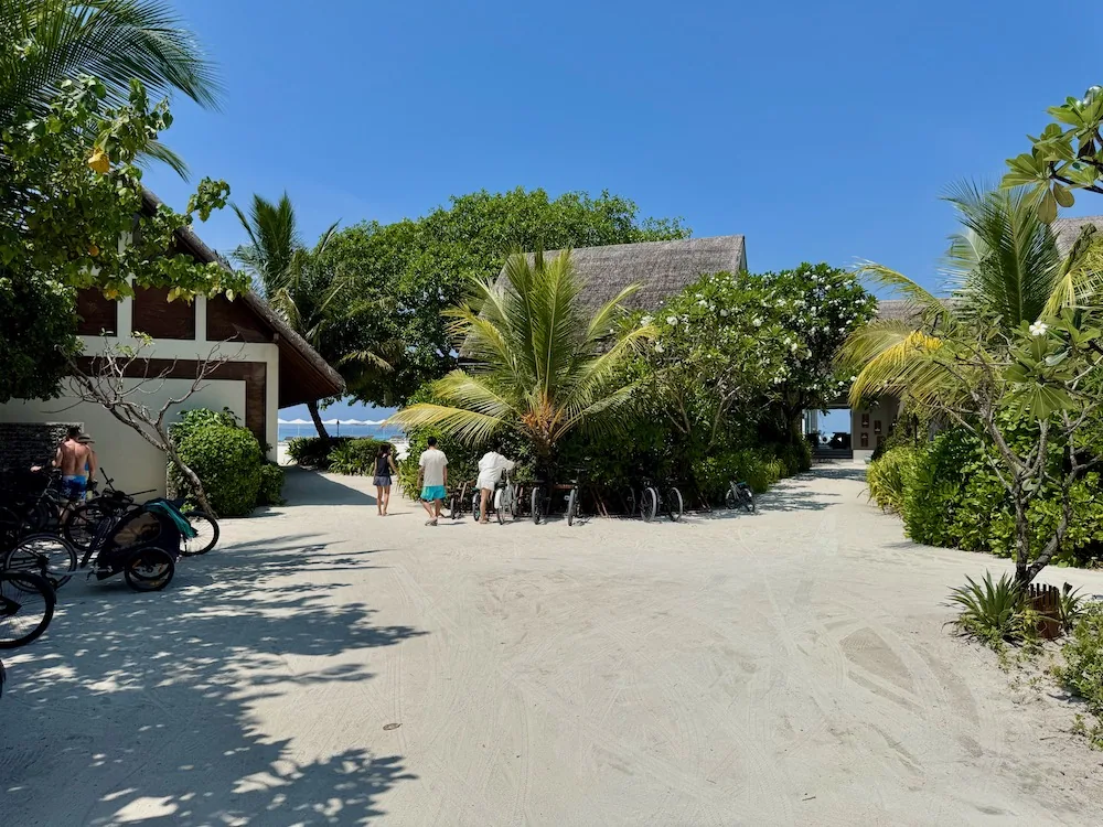 Four Seasons Resort Maldives at Landaa Giraavaru, club, Baa Atoll, Maldives, Blu Beach Club approach: bicycles parked along the sand path between the white-rendered beach club pavilions, families walking through the palm canopy toward the beachfront.
