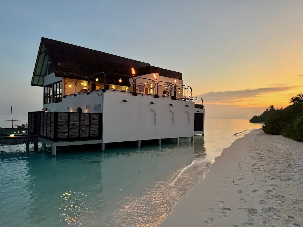Four Seasons Resort Maldives at Landaa Giraavaru, dining, Baa Atoll, Maldives, Al Barakat at sunset: a two-storey overwater dining pavilion with a row of Moroccan-arched windows along the white-rendered base, the upper terrace lit with hanging lanterns and torches, the beach curving along to the right.