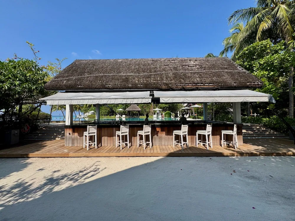 Four Seasons Resort Maldives at Landaa Giraavaru, pool, Baa Atoll, Maldives, Pool bar: thatched-roof structure with a row of seven white timber bar stools on the teak deck, white awnings extended either side, the main pool deck and daybeds visible behind.