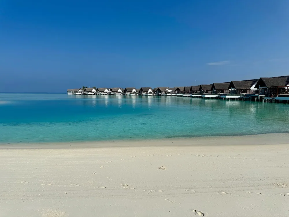 Sunset Water Bungalow row from the beach: a long curved line of identical thatched-pyramid roof bungalows on stilts marching across the lagoon, the western over-water arrangement seen at low water.