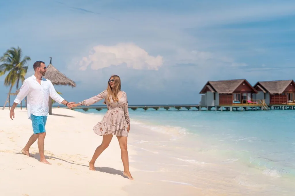 Furaveri Maldives, couple walking hand-in-hand on white-sand beach with thatched overwater villa cluster on right, palms and clear lagoon, Raa Atoll, Maldives