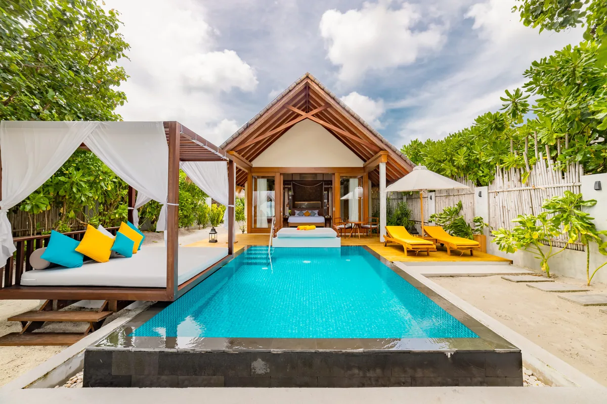Furaveri Maldives, Beach Pool Villa exterior with pitched-roof villa, infinity-edge blue-tile pool, draped four-poster daybed and yellow sun loungers under thatched parasol, Raa Atoll, Maldives