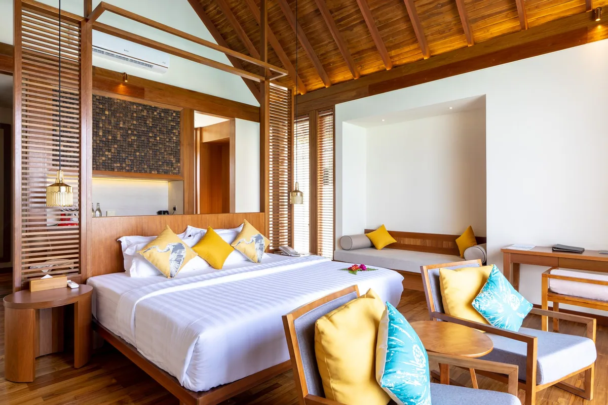 Furaveri Maldives, Ocean Pool Villa bedroom with cathedral wood-beam ceiling, dark wood louver dividers, king bed with yellow accents, banquette daybed and yellow-cushion armchairs, Raa Atoll, Maldives