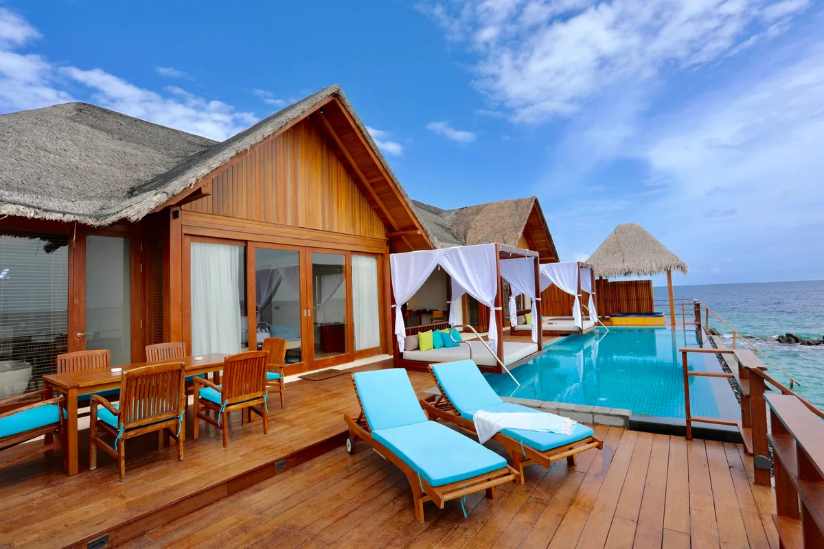 Furaveri Maldives, Sunset Pool Water Villa exterior with thatched roof, wood-panel exterior, infinity pool, draped four-poster daybed pavilion and sun loungers, Raa Atoll, Maldives