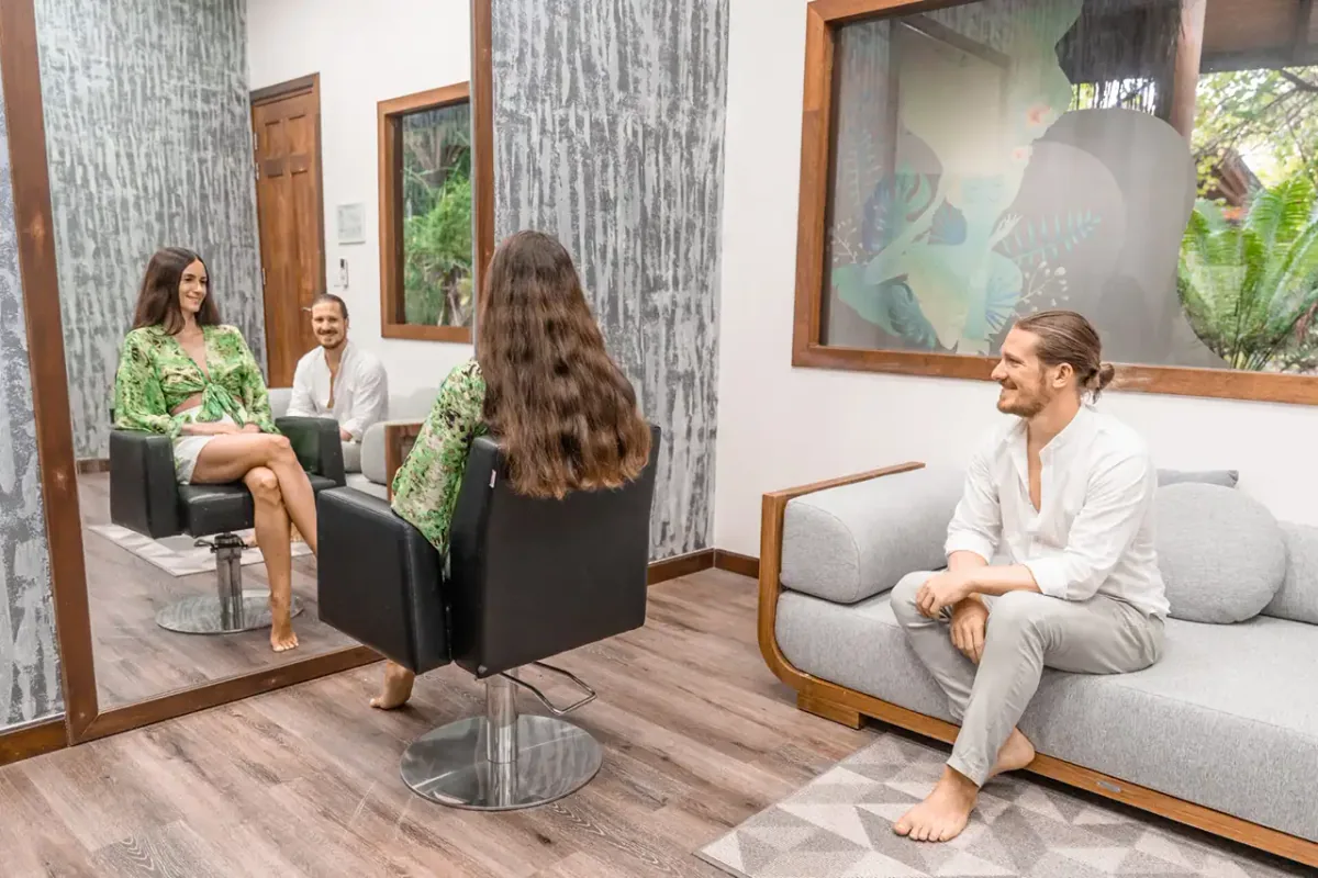 Fushifaru Maldives, spa, Lhaviyani Atoll, Maldives, Hair-and-beauty salon interior with a guest in the styling chair facing the mirror, a companion on the grey sofa, tropical-leaf wall art, and textured grey wall covering.