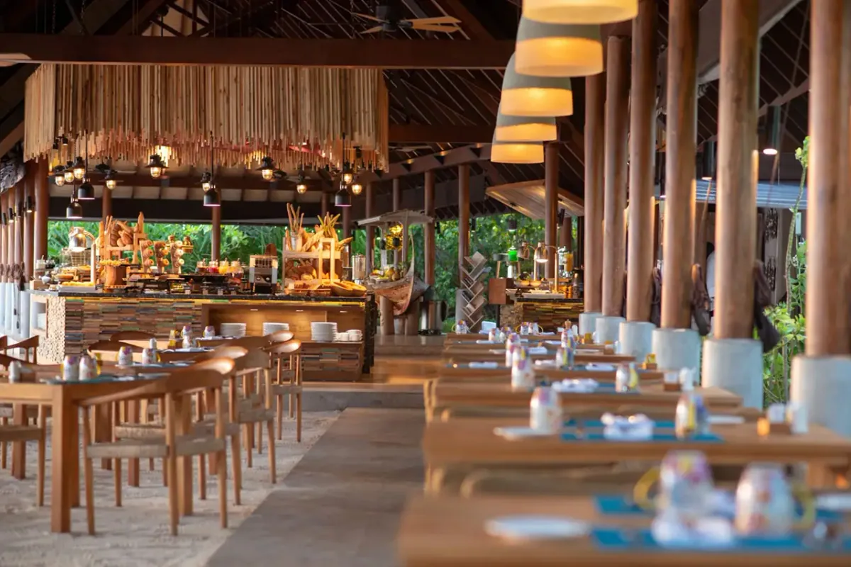 Fushifaru Maldives, dining, Lhaviyani Atoll, Maldives, Korakali all-day pavilion with vaulted timber rafters, a hanging woven-straw curtain installation overhead, conical fabric pendant lights, and a buffet station beyond the long wooden tables.