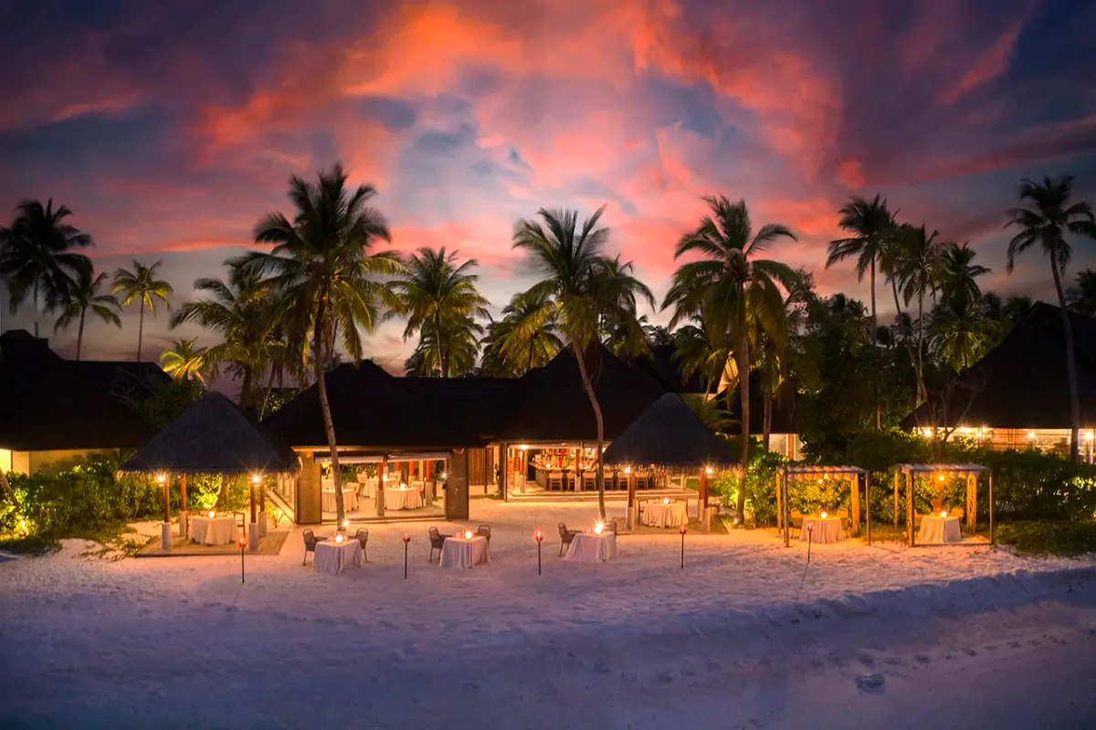 Fushifaru Maldives, dining, Lhaviyani Atoll, Maldives, Raakani fine-dining beach setup at sunset with thatched-cone pavilions, individual candle-lit tables on the sand, and palm silhouettes against a pink dusk sky.