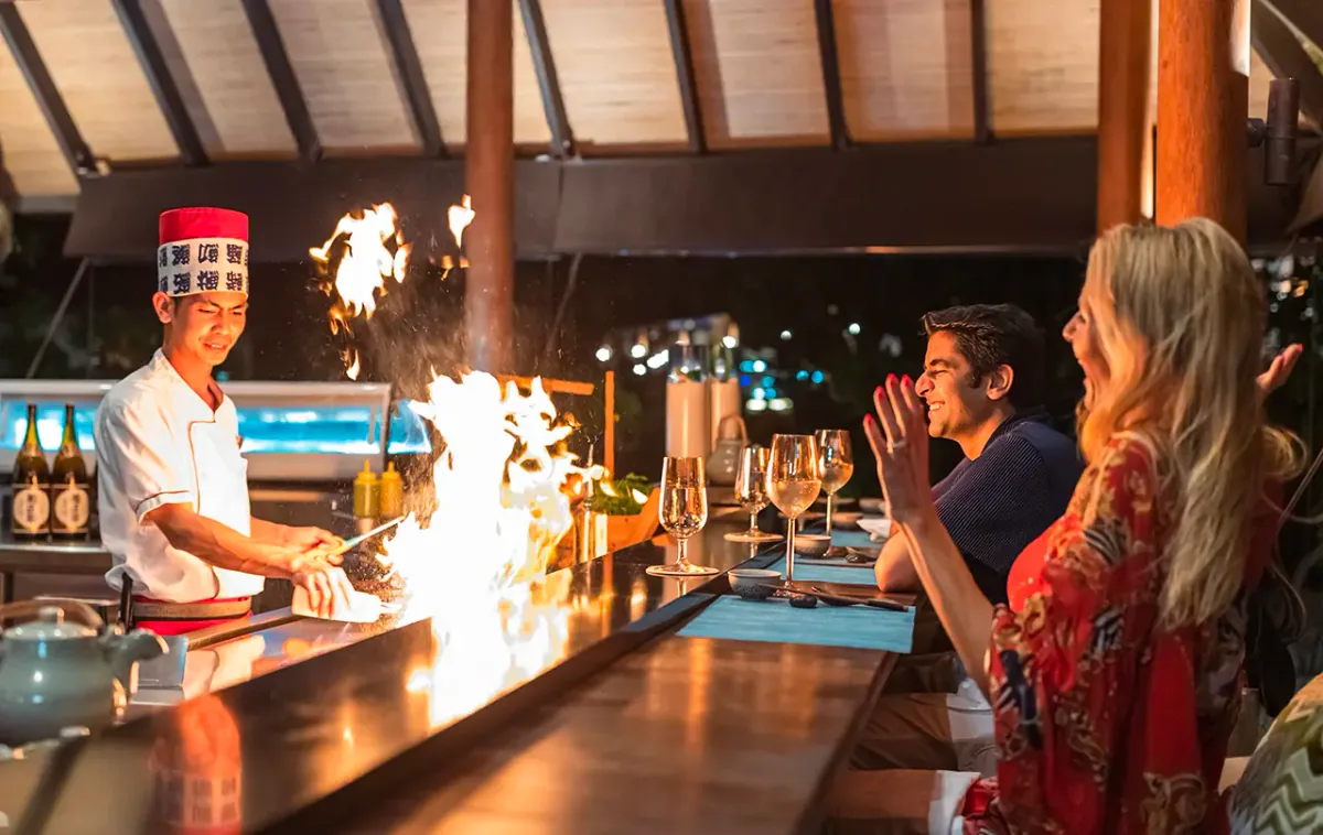 Fushifaru Maldives, dining, Lhaviyani Atoll, Maldives, Teppanyaki counter with the chef working a live flame, sake bottles on the back shelf, and two guests seated under a bamboo ceiling.
