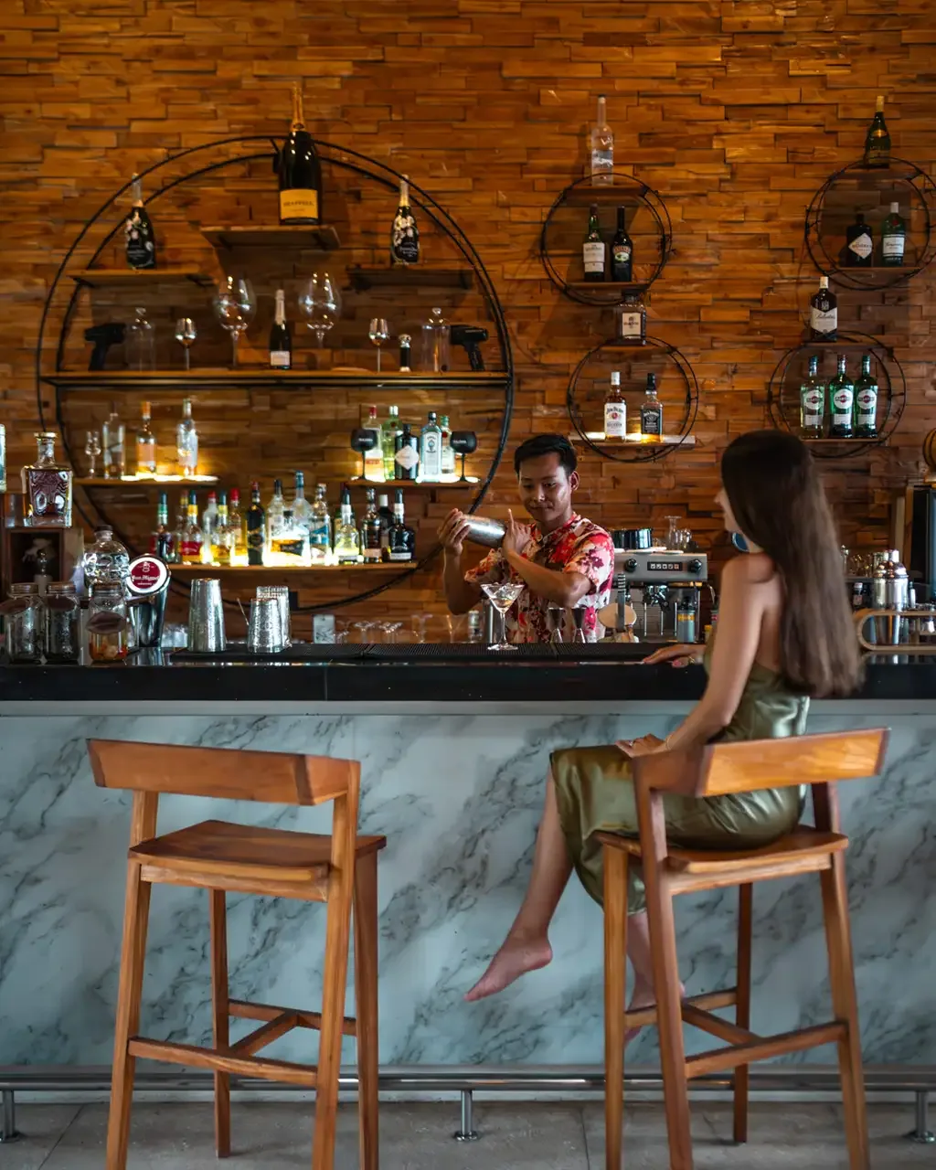 Fushifaru Maldives, dining, Lhaviyani Atoll, Maldives, Fanihandhi pool bar interior with a bartender shaking a cocktail behind a marble-clad bar, wood-shingled feature wall, and concentric metal-ring bottle shelves.