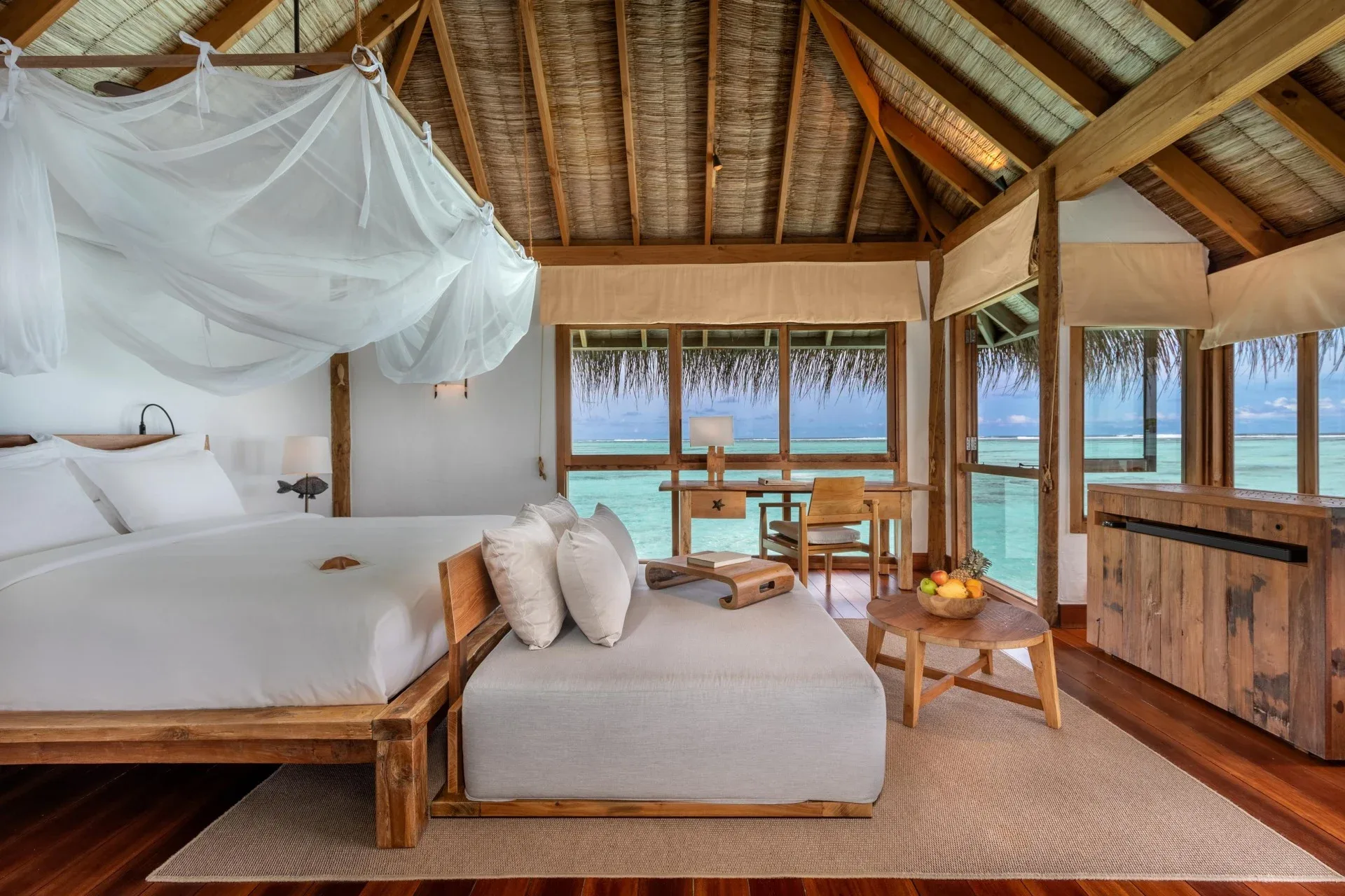 Gili Lankanfushi, villa overwater, North Malé Atoll, Maldives, Crusoe villa bedroom interior: vaulted thatched-roof ceiling with exposed timber rafters, king bed with white linens and a sheer canopy mosquito net, daybed ottoman, reclaimed-timber writing desk by the picture window with palm-frond canopy overhang, hardwood floors with woven jute mat.