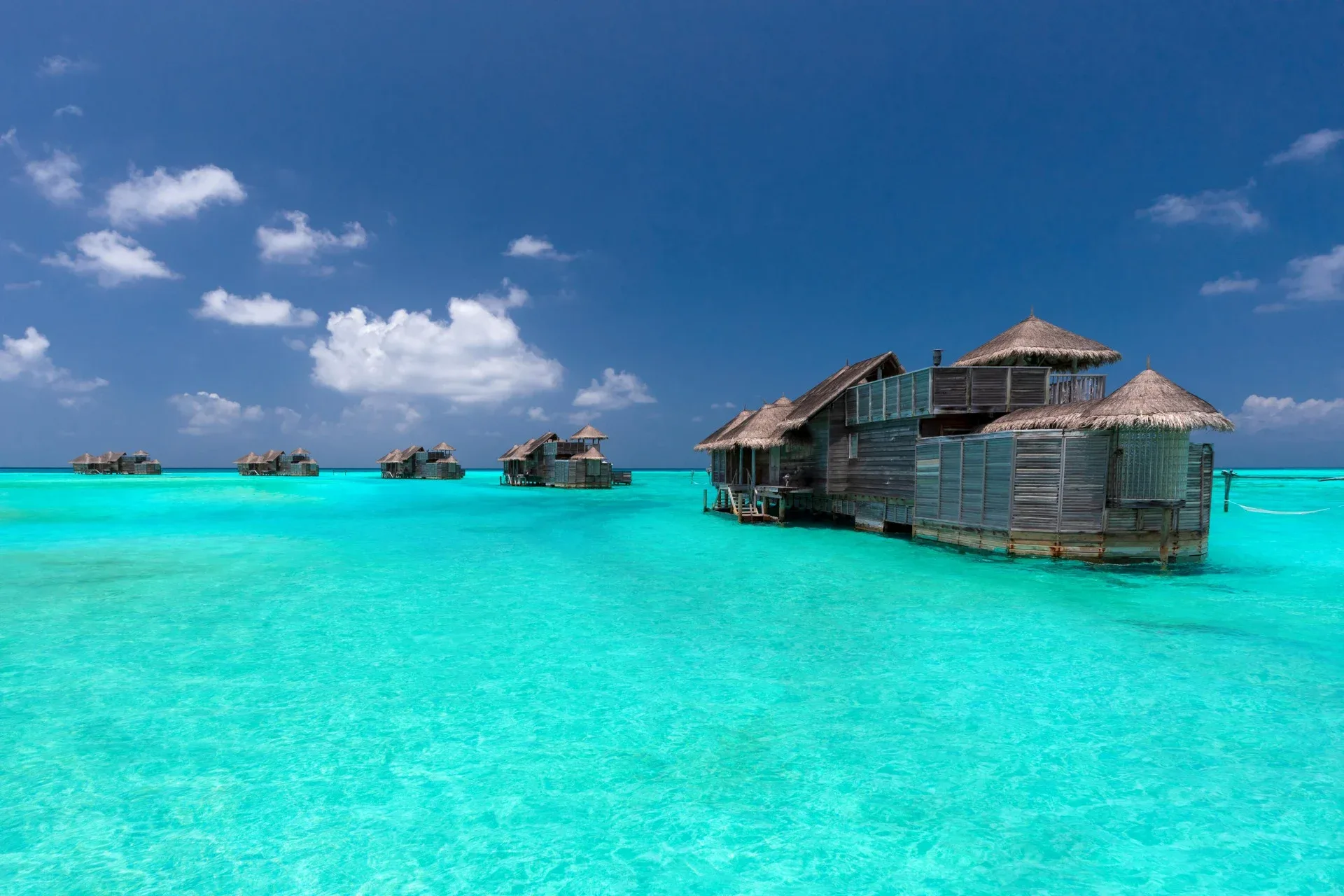 Gili Lankanfushi, villa overwater, North Malé Atoll, Maldives, The Crusoe Residences cluster, scattered standalone overwater pavilions sitting alone in the turquoise lagoon, the closest a two-storey multi-pod with weathered timber siding and twin thatched conical roofs.