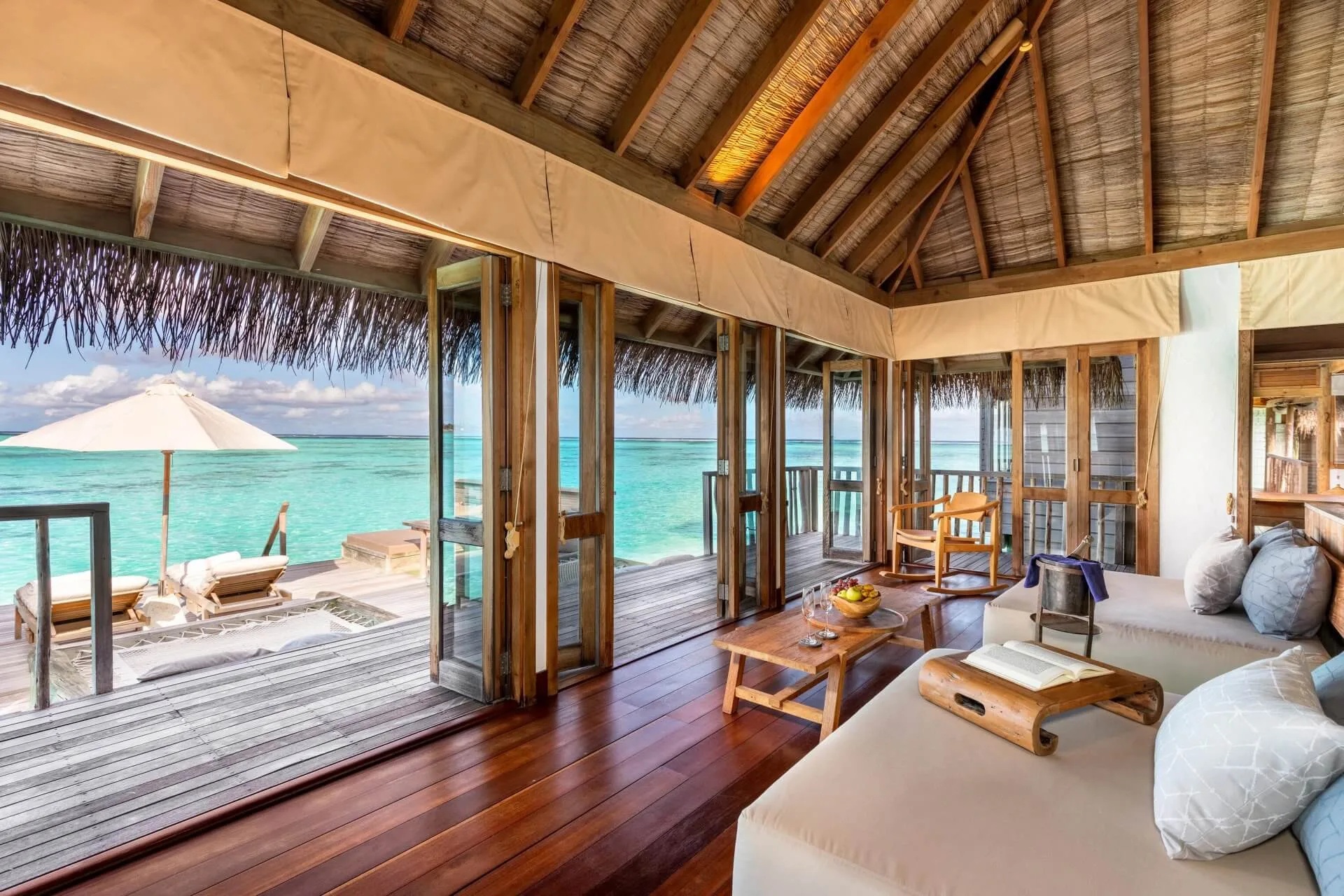 Gili Lankanfushi, villa overwater, North Malé Atoll, Maldives, Villa living pavilion: low timber-frame sofa with linen cushions, vaulted thatched ceiling, sliding doors open to the deck with sun loungers and lagoon view, neighbouring Crusoe-style overwater villas in the middle distance.
