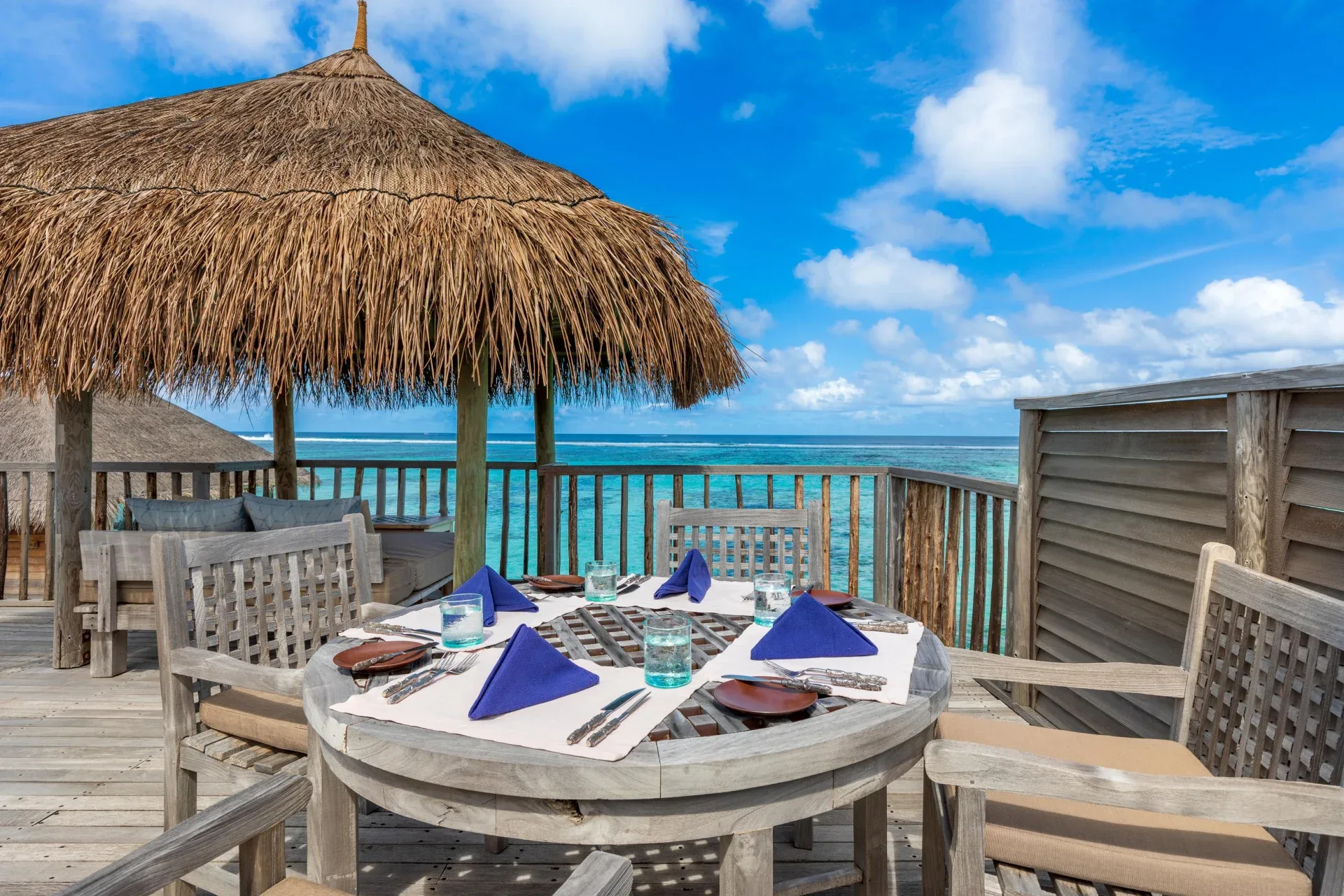 Gili Lankanfushi, villa overwater, North Malé Atoll, Maldives, Villa rooftop terrace dining: weathered timber dining table set for four under a thatched parasol, lagoon horizon, the rooftop space used for private breakfasts and sundowner setups.