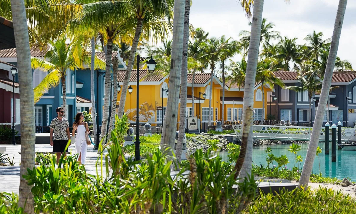 Hard Rock Hotel Maldives, setting, South Malé Atoll, Maldives, The Marina commercial precinct: a row of warehouse-style buildings in saturated yellow, teal, and navy with red-tile roofs, palm-lined promenade, a couple walking past the marina inlet, the retail-and-dining strip that makes Crossroads the country's only multi-property commercial precinct.