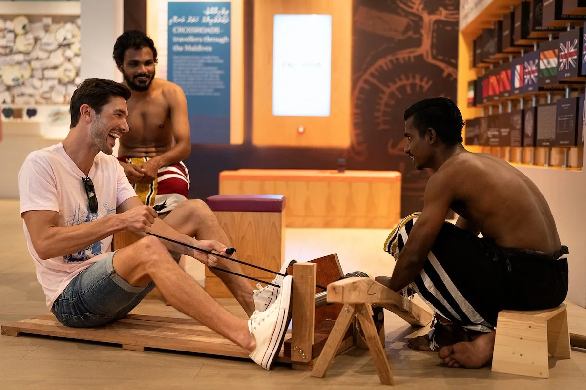 Hard Rock Hotel Maldives, activity, South Malé Atoll, Maldives, Cultural workshop at the Maldives Discovery Centre inside the Crossroads precinct: a guest seated on a wooden floor learning traditional Maldivian rope-and-weaving technique from two artisans, the CROSSROADS Travellers Through the Maldives exhibit signage and ancient Maldivian-script display in the background.