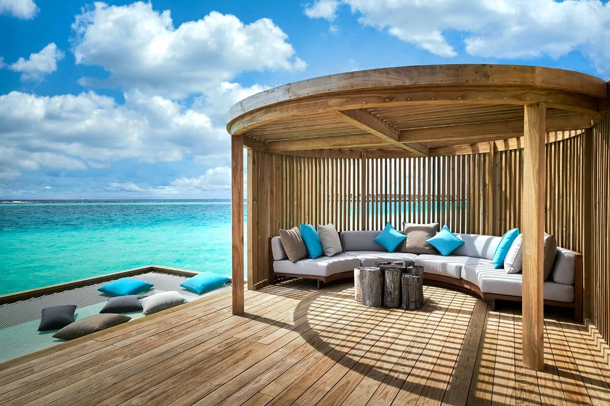 Hard Rock Hotel Maldives, setting, South Malé Atoll, Maldives, Overwater lounge cabana: a curved vertical-slat wooden pavilion on a teak deck, white sofas with teal pillows, log-stump side tables, a netted swing/lounger over the lagoon with cushions, the overwater villa cluster glowing on the horizon.