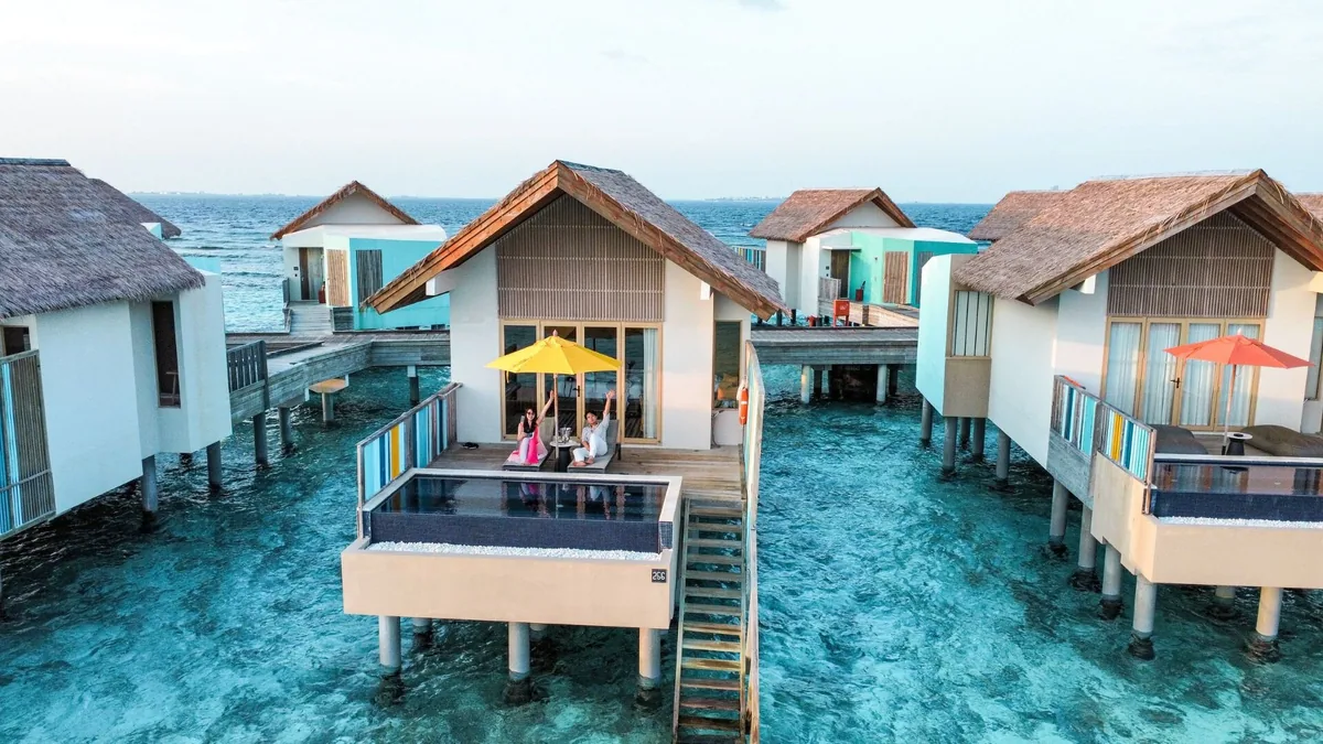 Hard Rock Hotel Maldives, aerial, South Malé Atoll, Maldives, Close-aerial of the overwater villa cluster: peaked thatched-roof villas standing on stilts, each deck carrying the signature multicolor louvered railing and a plunge pool with steps down to the lagoon, yellow and red parasols, couple visible on the closest villa deck.