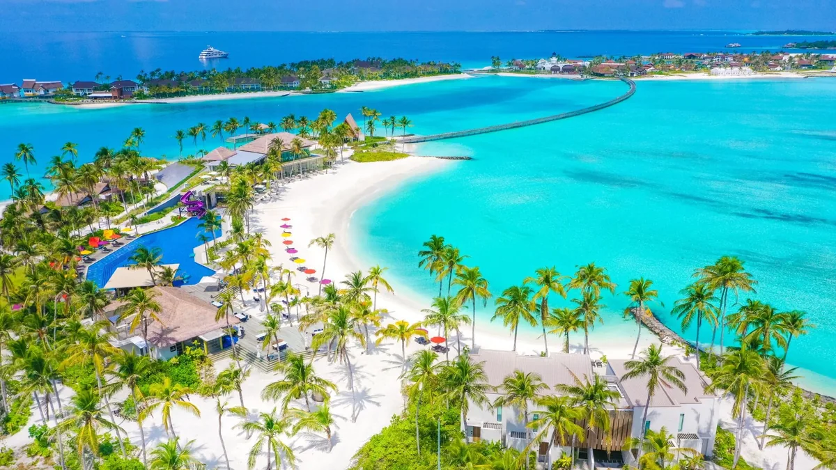 Hard Rock Hotel Maldives, aerial, South Malé Atoll, Maldives, Wide aerial of the Crossroads Maldives integrated-development cluster: Hard Rock anchor in the foreground (palm-dense island, pool, multicolor beach parasols), the long wooden boardwalk reaching out to the adjacent SAii Lagoon island, the Marina commercial precinct beyond, the country's only multi-property cluster geometry in a single frame.