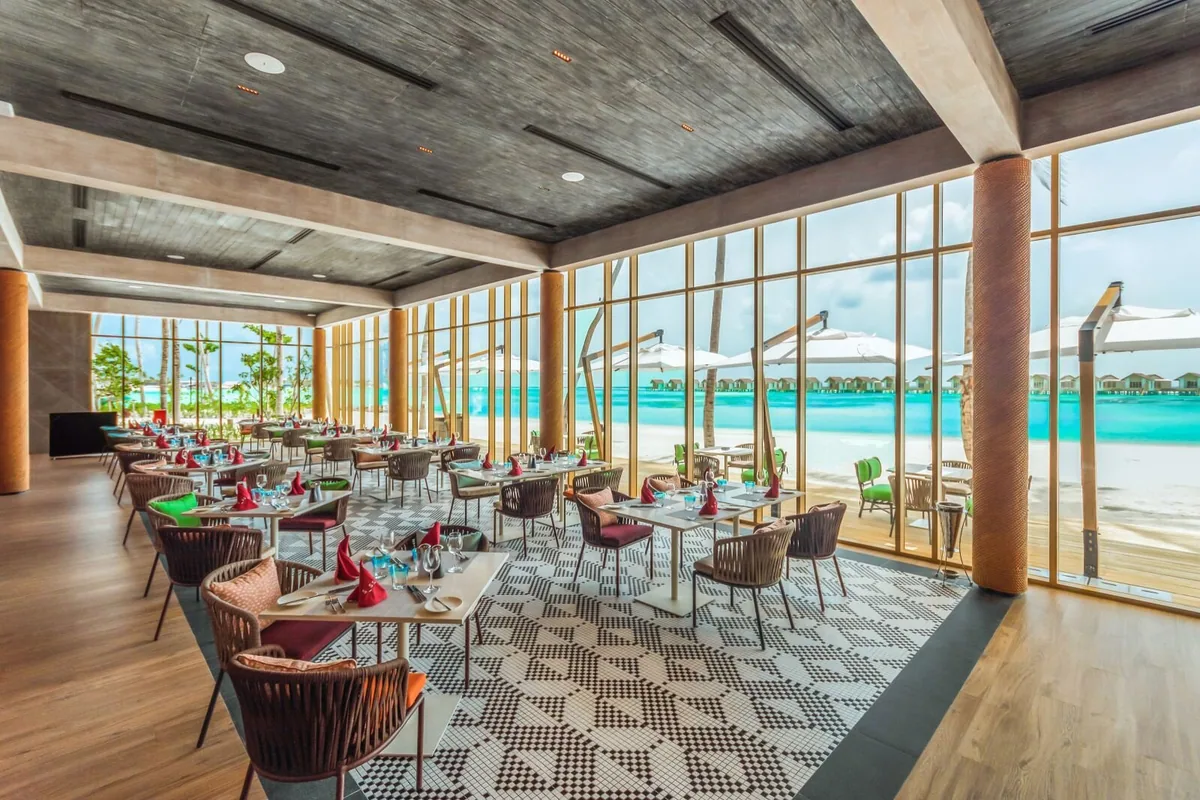Hard Rock Hotel Maldives, dining, South Malé Atoll, Maldives, Sessions principal dining pavilion interior: exposed concrete ceiling, wooden columns, geometric black-and-white tile floor, woven leatherette chairs at white-top tables, floor-to-ceiling windows opening onto the lagoon and the overwater villa cluster across the water.