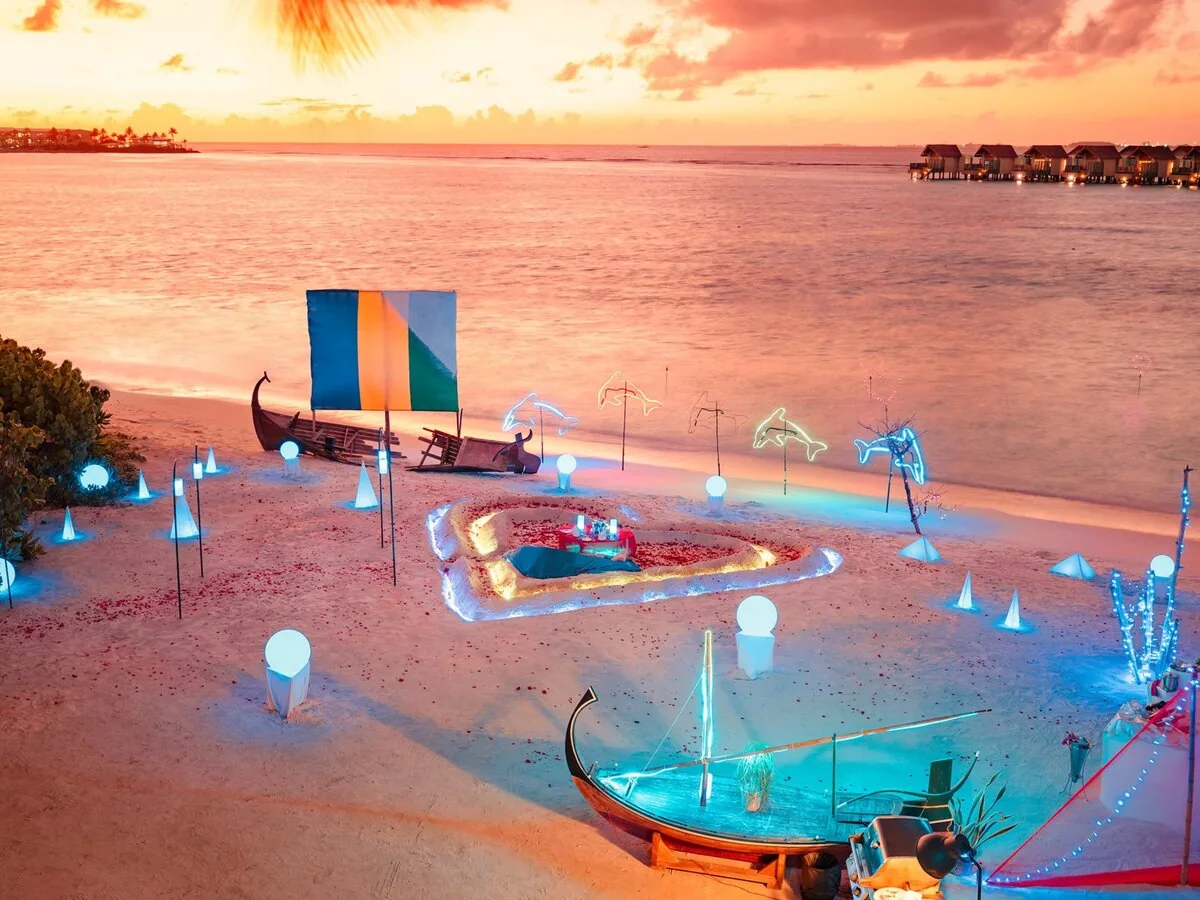 Hard Rock Hotel Maldives, dining, South Malé Atoll, Maldives, Sunset beach celebration setup: a candle-lit private dinner table inside a heart-shaped sand pit ringed with rose petals, a traditional Maldivian dhoni boat decorated with multi-color string lights, neon dolphin-and-music-note sculptures, sail flying the Hard Rock blue-yellow-green colors, bulb-stake lights along the shore, the overwater villa cluster glowing across the lagoon at twilight.