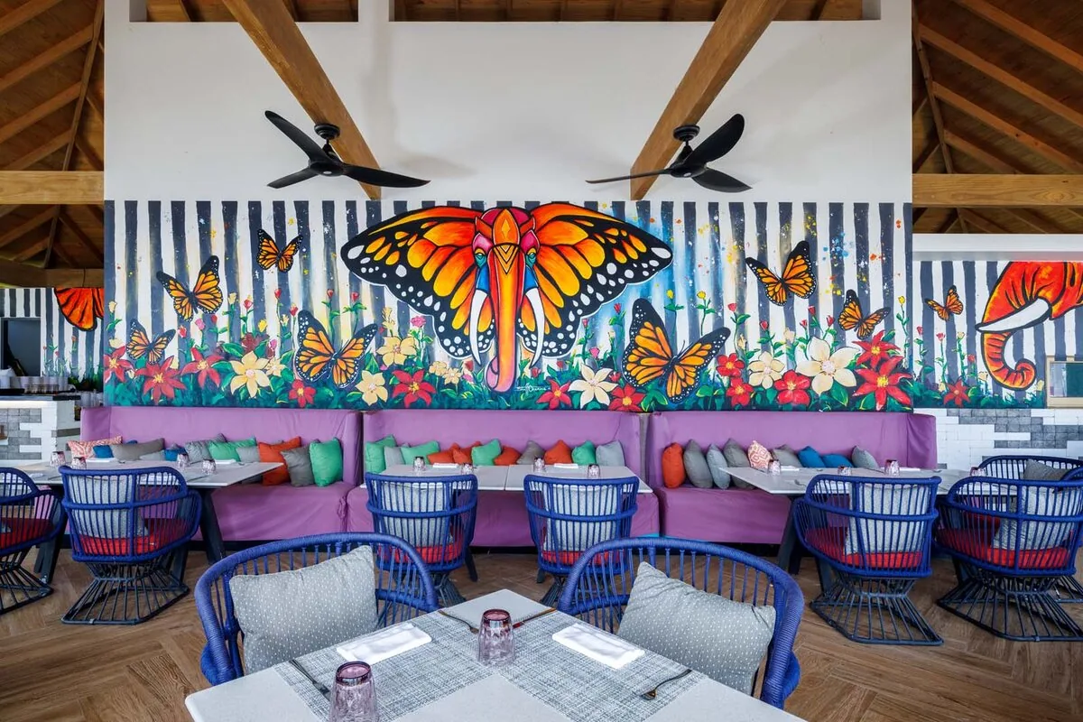 Hard Rock Hotel Maldives, dining, South Malé Atoll, Maldives, Elephant and Butterfly venue: vibrant monarch-butterfly-and-floral mural feature wall against navy stripes, exposed wooden ceiling beams, navy-blue rattan barrel chairs at marble-top tables, purple banquette seating with multicolor accent pillows, the second-venue counterpoint to the Sessions main pavilion.