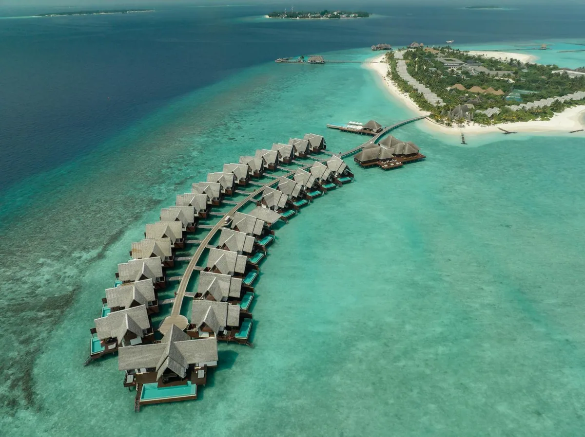Heritance Aarah Maldives, setting, Raa Atoll, Maldives, Arced overwater villa row aerial showing 25+ A-frame thatched-roof villas with cantilevered green-tile plunge pools along the eastern Aarah reef, the main island beach and dining cluster anchoring the right-hand frame (Heritance Aarah signature shot).
