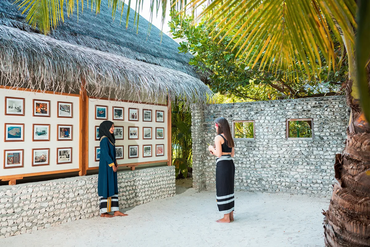 Heritance Aarah Maldives, lifestyle, Raa Atoll, Maldives, The Maldivian cultural exhibit pavilion, stone-walled enclosure with thatched-roof entry housing framed historical photographs, two figures (one in traditional libaas dress) examining the display in soft midday light.