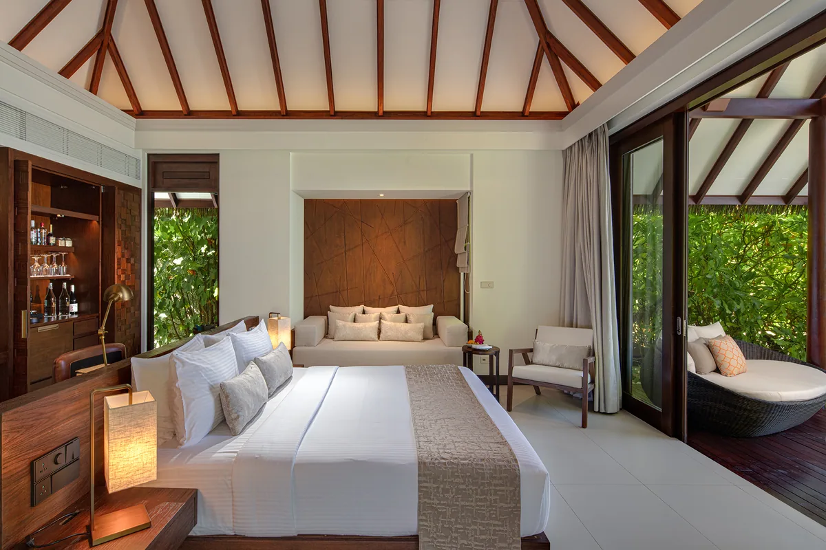 Heritance Aarah Maldives, villa beach, Raa Atoll, Maldives, Heritance Beach Villa bedroom interior with exposed wooden roof trusses under the A-frame thatched ceiling, king bed with bronze pendant lamps, daybed sofa, mosaic-accent minibar console, sliding glass doors to garden, woven outdoor recliner.