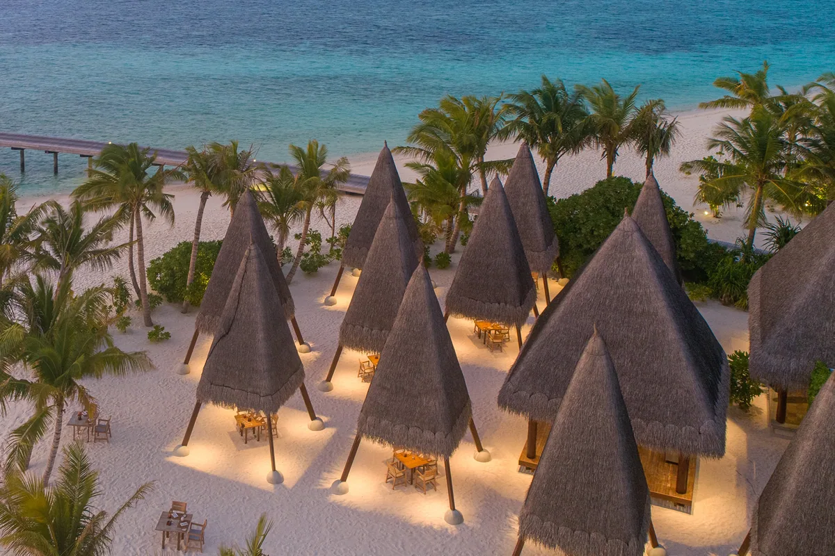 Heritance Aarah Maldives, dining, Raa Atoll, Maldives, Ginifati private dining beach cluster at twilight, cone-shaped thatched-roof pavilions arranged along the white-sand beach with each table set for two, palm trees framing, soft amber pavilion lighting.