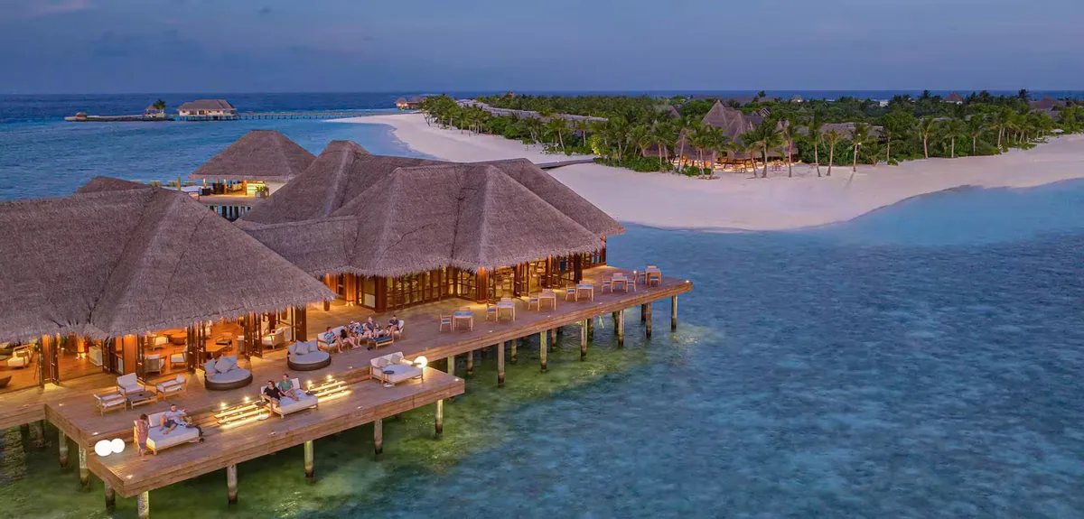 Heritance Aarah Maldives, dining, Raa Atoll, Maldives, Overwater dining cluster at twilight, three connected thatched-roof restaurant pavilions (Ralu, Ambula, Hathaa overwater venues) extending on a deck above the lagoon with lit lounge daybeds at deck level and the main island beach and tree line in the background.