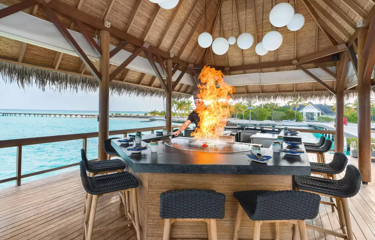 Heritance Aarah Maldives, dining, Raa Atoll, Maldives, Hathaa overwater Japanese teppanyaki counter, chef performing the live-flame fire show across the polished black grill, woven black bar chairs around the counter, spherical white pendant lights under the thatched pavilion, the OWV row and main island reading across the open lagoon.
