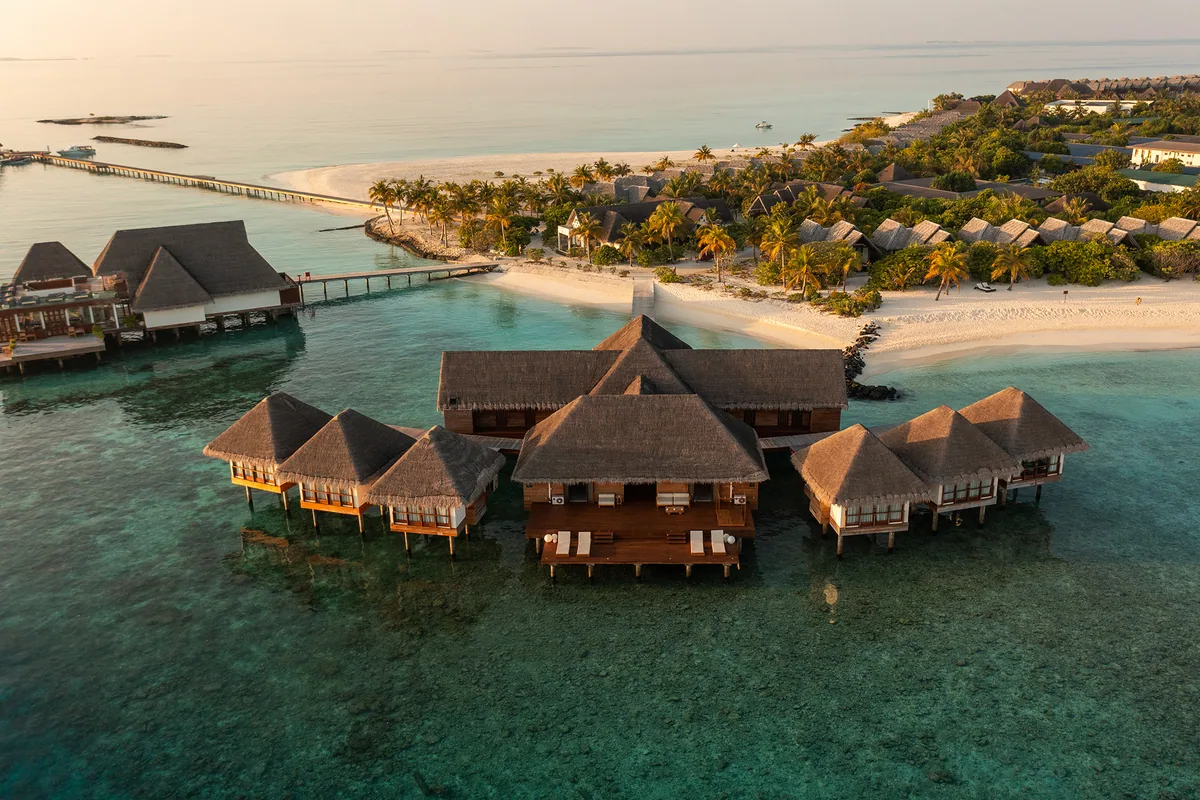 Heritance Aarah Maldives, spa, Raa Atoll, Maldives, IASO Spa aerial showing the six thatched-roof overwater treatment pavilions clustered above the lagoon, connected by timber bridges to the spa reception and beach beyond, dusk light catching the thatch.