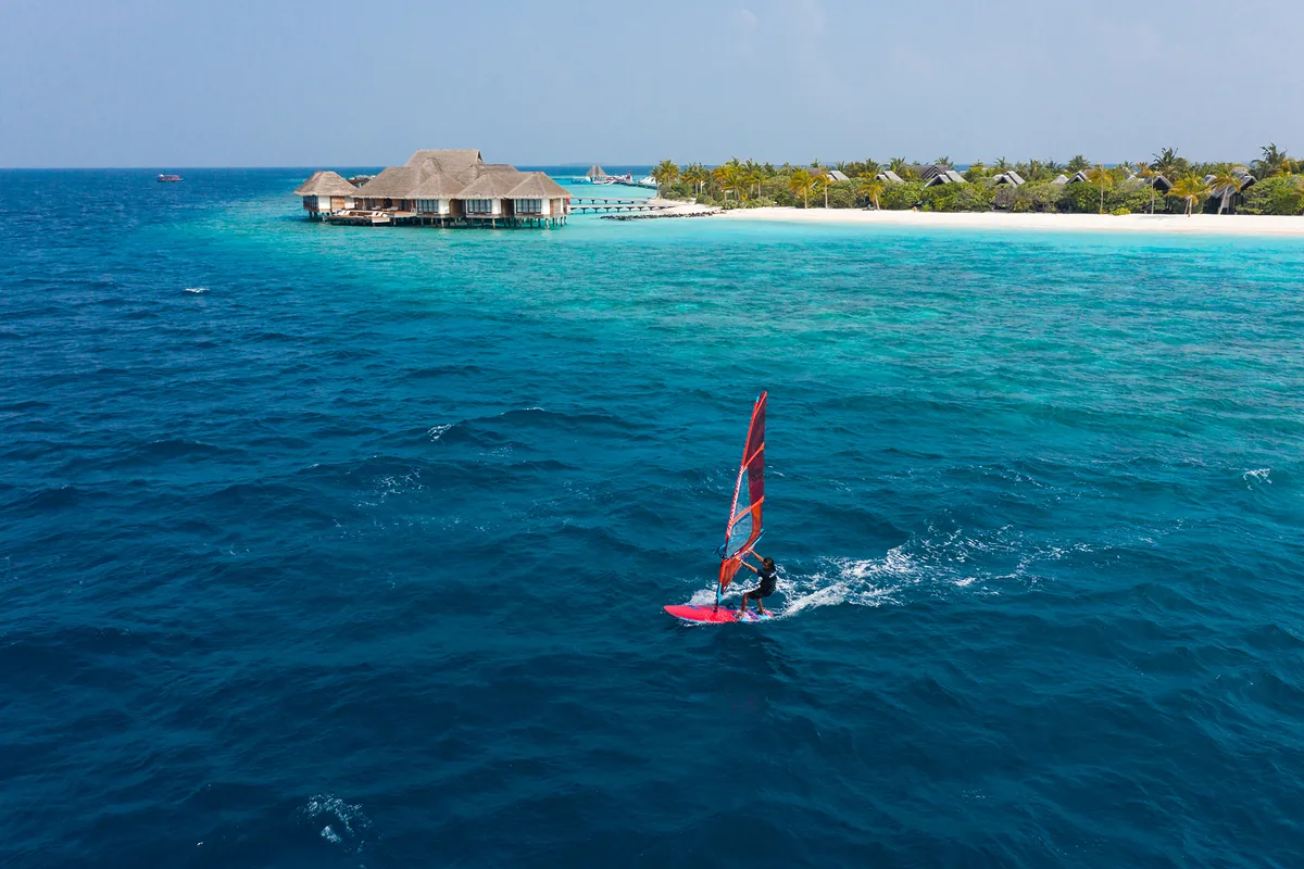 Heritance Aarah Maldives, activities, Raa Atoll, Maldives, Windsurfer crossing the deep-blue Aarah lagoon under a red sail, the overwater pavilion arm and beach-villa row reading distinctly along the right-hand frame.