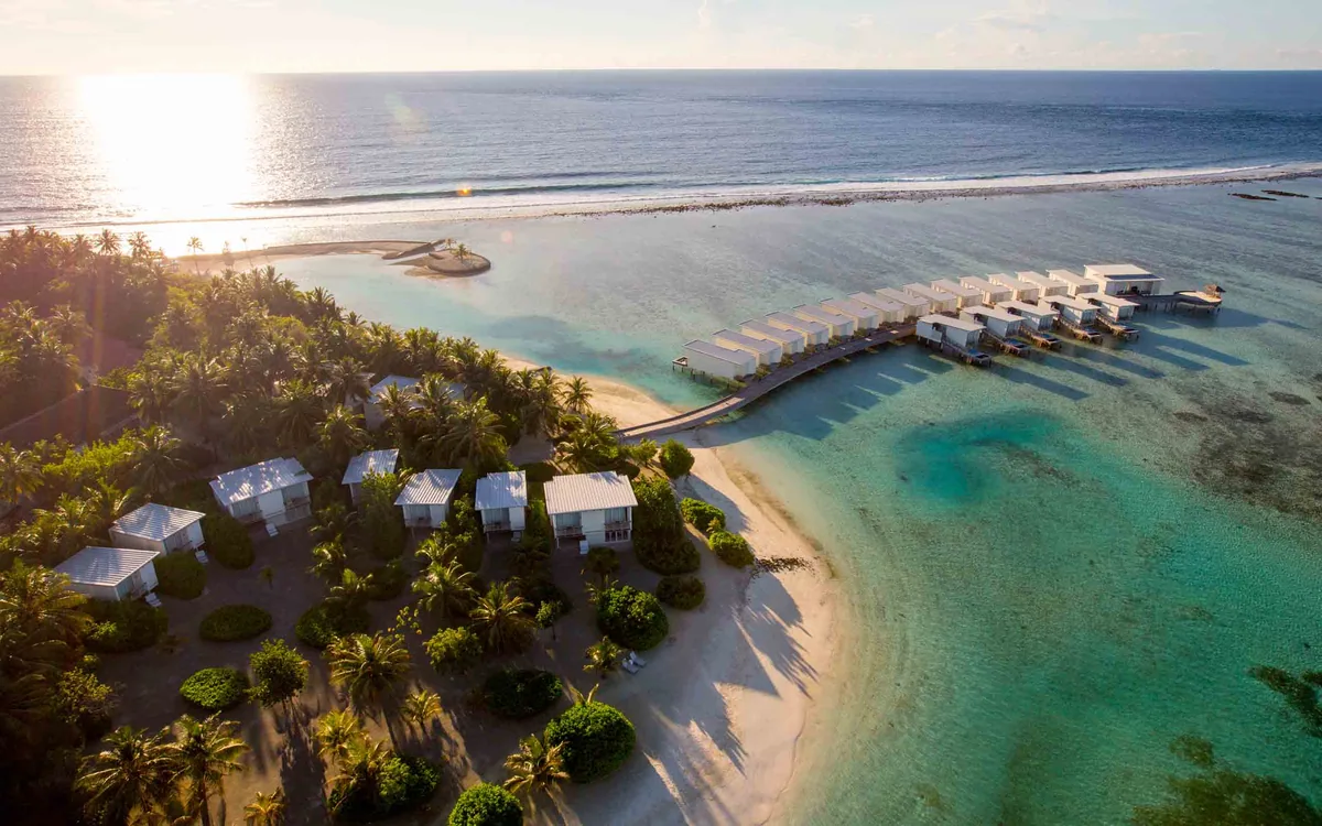 Holiday Inn Resort Kandooma Maldives, aerial, South Malé Atoll, Maldives, Sunrise aerial of Holiday Inn Kandooma: white flat-roof beach villas in the palm-fringed foreground, the long boardwalk reaching out to the over-water villa cluster on the right, the Kandooma Right reef break visibly peeling on the horizon at the property's southern edge, golden sunrise light raking across the lagoon.