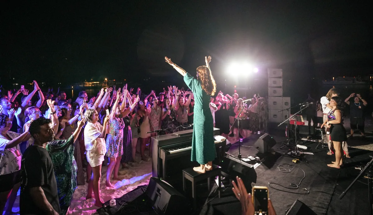 Holiday Inn Resort Kandooma Maldives, lifestyle, South Malé Atoll, Maldives, Beach concert at night under stage lighting: a singer on a low stage with arms raised, the crowd of guests packed onto the sand with arms up and phones held out, keyboard and amp stack flanking the stage, the documentary face of the Kandooma 2026 concert series that runs multiple sold-out weeks each year.