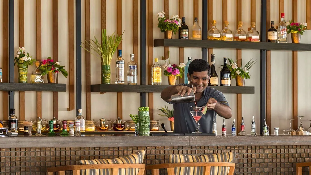 Hurawalhi Island Resort, dining, Lhaviyani Atoll, Maldives, Kashibo bar with a bartender pouring a martini, a vertical wood-slat back wall with floral arrangements and bottles displayed on black-frame shelves, and a mosaic bar front.