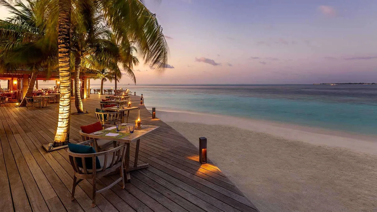 Hurawalhi Island Resort, dining, Lhaviyani Atoll, Maldives, Canneli beach-deck dinner setting at dusk with a row of wooden tables and chairs along the deck edge, candle lanterns, warm-lit palm trunks, and the lagoon at the right.