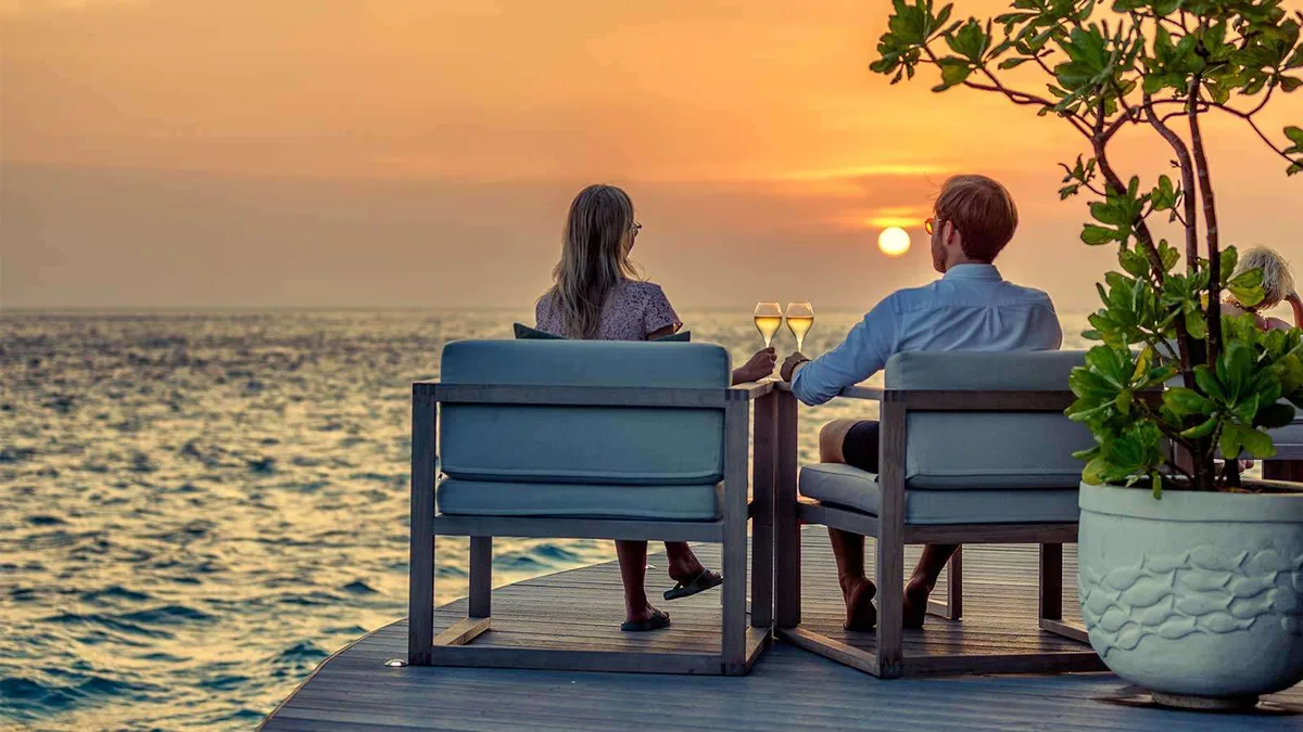 Hurawalhi Island Resort, dining, Lhaviyani Atoll, Maldives, Champagne Pavilion overwater deck at sunset with two blue cushioned armchairs and a couple lifting champagne flutes against the dusk horizon.