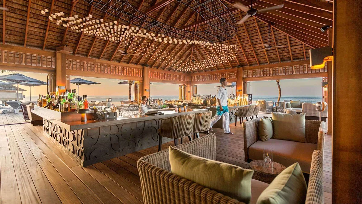 Hurawalhi Island Resort, dining, Lhaviyani Atoll, Maldives, Coco bar with an open thatched pavilion roof, a cascade of cluster pendant lights overhead, a long bar with decorative metalwork front, wicker armchairs in the foreground, and sunset light beyond the open sides.