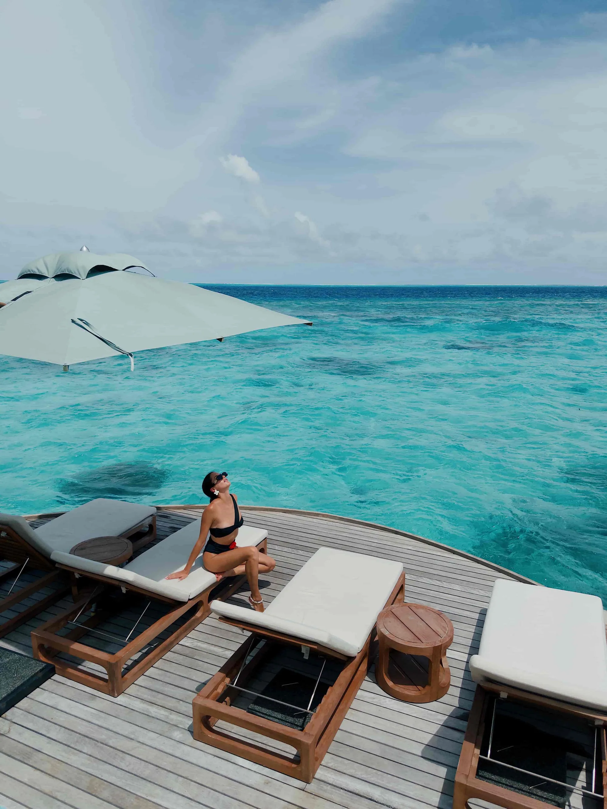 Huvafen Fushi, arrival, North Malé Atoll, Maldives, Overwater sun-deck arrival reading, beige cushioned daybeds and sun loungers under a canvas parasol on the timber deck, a guest perched at the deck edge looking out over the open lagoon toward the reef break.